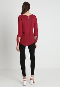 Blusa bordeaux con maniche a tre quarti e zip sul retro, abbinata a leggings neri e sandali bianchi con tacco a blocco. Tessuto morbido, vestibilità casual.