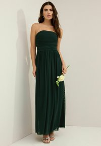 Off-shoulder maxiklänning i mörkgrön tyg med rynkad byst och flödande kjol. Modellen håller en vit ros med grönska och bär utsmyckade sandaler.