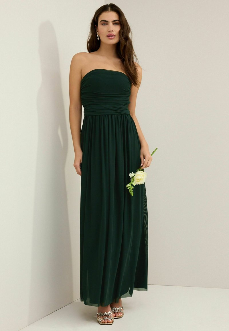 Off-shoulder maxiklänning i mörkgrön tyg med rynkad byst och flödande kjol. Modellen håller en vit ros med grönska och bär utsmyckade sandaler.