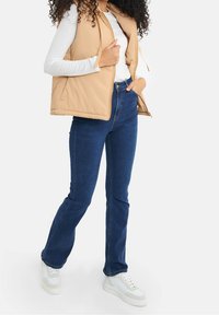 Beige gepolsterte Weste mit hohem Kragen, mit durchgehendem Reißverschluss, getragen über einem weißen Langarmshirt, kombiniert mit dunkelblauen Schlagjeans und weißen Sneakers.