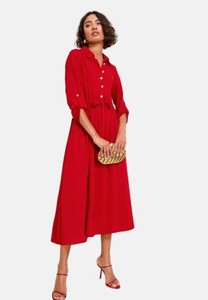 Woman wearing a red long-sleeve midi dress with buttons, holding a gold clutch, and wearing red high-heeled sandals, standing on a plain background.