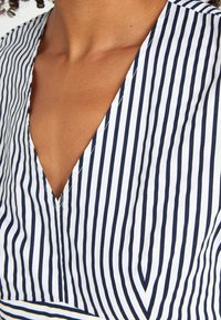 Blouse rayée avec un décolleté en V profond. Présente des rayures verticales étroites bleues marines et blanches, un tissu lisse et une coupe décontractée.