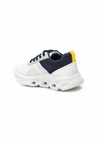 Zapatilla deportiva blanca y navy con parte superior de malla, lengüeta acolchada y acentos texturizados. Presenta una suela blanca gruesa con recortes. Lengüeta trasera amarilla.