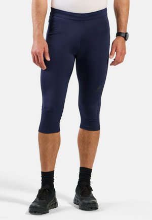 Man wearing navy blue knee-length athletic leggings, black socks, black running shoes, white shirt, and black smartwatch on left wrist.