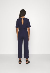 Marineblaues Jumpsuit mit kurzen Ärmeln, Schlüsselöffnung im Rücken, strukturiertem Stoff und weiten, verkürzten Beinen. Getragen mit schwarzen Riemchensandalen.