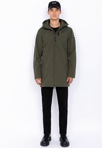 Olijfgroene waterdichte jas met een ritssluiting aan de voorkant, verstelbare capuchon en twee zijzakken, gedragen over een zwarte broek en laarzen.