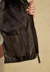 Brūna quiltota jaka ar gludu tekstūru iekšpusē, ar kabatu un izsmalcinātām iepresētām zīmēm. Zibens aizdare apakšā.