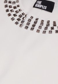 Biała swetra z okrągłym dekoltem, zdobiona srebrnymi piramidalnymi ćwiekami wokół dekoltu. Metka głosi "The Kooples". Miękka faktura z prążkowanej tkaniny.