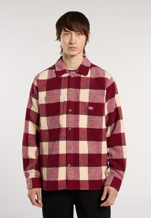 Joven vestido con una camisa de cuadros grande en rojo y crema, con dos bolsillos en el pecho y pantalones negros, de pie contra un fondo liso.
