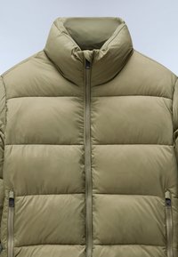 Olivgrön puffjacka med hög krage, horisontellt quiltad design och en framzipper; har sidofickor med dragkedjestängning.
