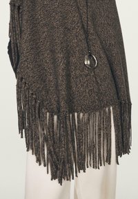 Cardigan marrone lavorato a maglia con dettagli di frange all'orlo e ai lati. Presenta un elemento decorativo circolare e un tessuto strutturato.