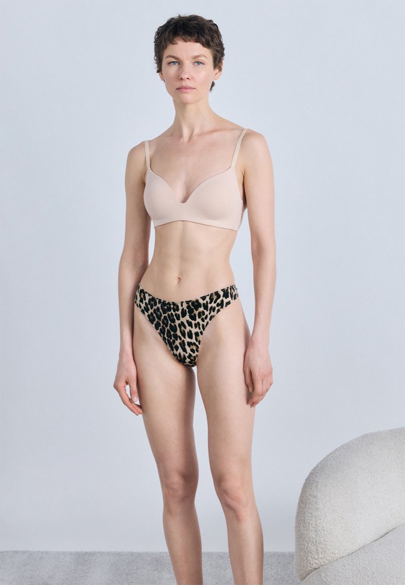 Femme aux cheveux courts portant un soutien-gorge beige et une culotte imprimé léopard, debout à côté d'un fauteuil blanc texturé sur un fond clair.