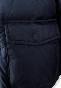Section d'une veste matelassée bleu marine avec un tissu texturé, une grande poche à rabat et un logo rond sur le rabat de la poche.