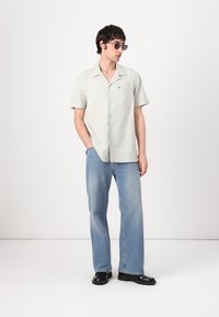 Hellgrünes, kurzärmeliges Hemd mit einer Brusttasche, kombiniert mit verfärbten blauen Weitbein-Jeans und schwarzen Schuhen. Das Modell trägt runde Sonnenbrillen.