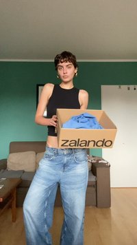Kartono dėžė su "Zalando" logotipu, kuri viduje turi mėlyną drabužį. Modelis dėvi juodą be rankovių marškinėlį ir šviesius platėjančius džinsus.