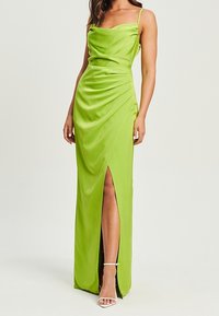 Kobieta ubrana w limonkową satynową sukienkę na ramiączkach z rozcięciem do uda i białe sandały na kostkę, stojąca na jednolitym tle.