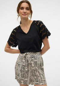 Camiseta negra con encaje, mangas en forma de onda y escote en V, combinada con pantalones cortos beige que tienen un estampado abstracto negro y una cinturilla atada.
