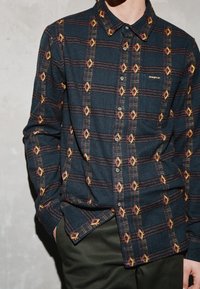 Mies, joka pukeutunut tummansiniseen ruudulliseen nappipaitaan, jossa keltaisia ja oransseja timantinmuotoisia kuvioita, toinen käsi taskussa, seisoo vaaleaa seinää vasten.