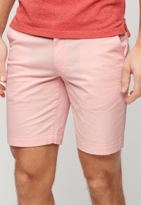 Homme portant un short rose clair ajusté et une chemise rouge, debout avec les mains détendues sur les côtés, sur un fond neutre.