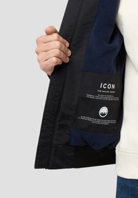 Schwarze Seemannsjacke mit navy-blauem Innenfutter, ausgestattet mit einem gesteppten Textur und einem Etikett mit Marken- und Nachhaltigkeitsinformationen.