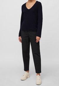 Persoon in een marineblauwe V-hals gebreide trui, zwarte rechte broek en crème chunky veterschoenen, staande voor een eenvoudige lichtgrijze achtergrond.