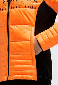 Puffer jacket features bright orange nylon material with stitched panels, a zippered front, and black fabric accents on the sides and sleeves.