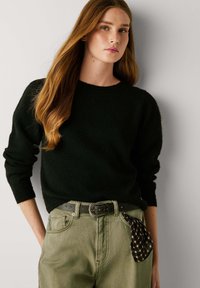 Melnas vilnas džemperis ar garām piedurknēm, apaļa izgriezuma kakls, kombinēts ar olīvu zaļām biksēm. Ievērojama sudraba jostas sprādze un melns šaha laukuma raksta kabatas lakats.