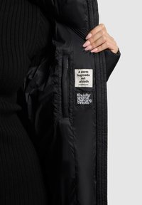 Melns puffersjakts ar gludu, spīdīgu tekstūru. Iekšpusē ir divi uzlīmes: viena ar tekstu krēmkrāsā fona, otra ar melnu diegu.