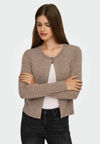 Cárdigan marrón de canalé con mangas largas, cuello redondo y corte ajustado, combinado con una blusa blanca y jeans oscuros, que destacan por un detalle de botones.