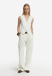 Gilet bianco in lino con scollatura profonda a V e bottoni frontali, abbinato a pantaloni bianchi larghi. Presenta una vita larga e nessun motivo visibile.