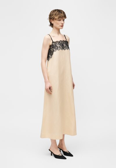 Γυναίκα που στέκεται, φοράει μπεζ αμάνικο φόρεμα με μαύρη δαντέλα στο στήθος και μαύρες μυτερές γόβες.