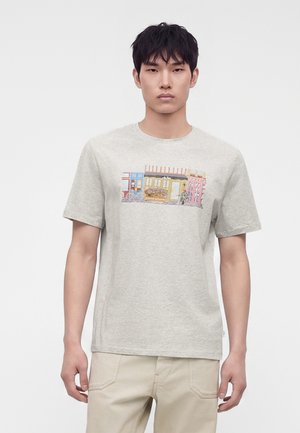 Jeune homme portant un t-shirt gris clair avec un motif coloré représentant une rue de la ville et un pantalon beige, se tenant devant un fond blanc uni.
