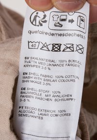 Etichetta di cura in tessuto bianco, contenente le istruzioni di cura in più lingue, con icone per il lavaggio e l'asciugatura. Superficie texturizzata.