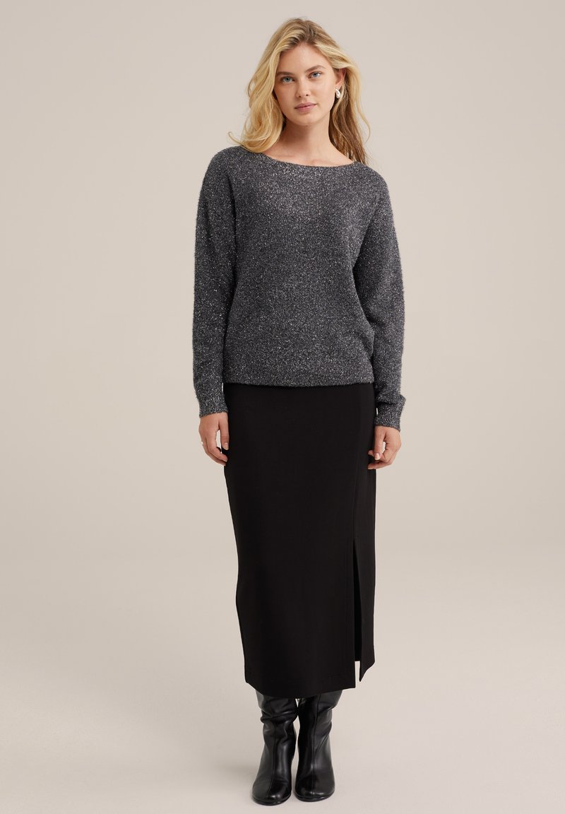 Pull en tricot gris à manches longues, col bateau, associé à une jupe midi noire avec une fente sur le côté, et des cuissardes noires.