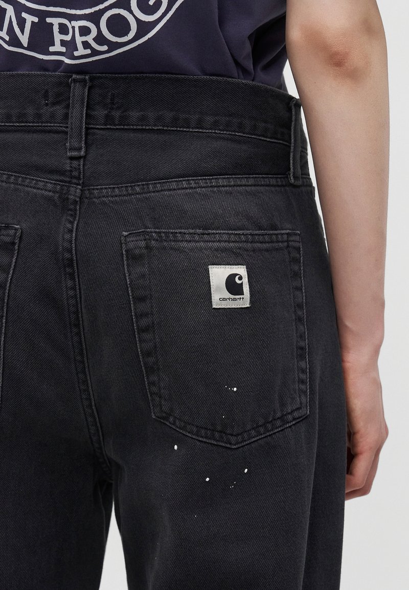 Jeans in denim nero con schizzi di vernice bianca sull'area della tasca posteriore e una toppa con il logo Carhartt sopra la tasca, indossati da una persona.