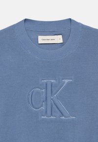Μπλε βαμβακερή μπλούζα με ribbed κολάρο, με κεντημένο λογότυπο "CK" σε αντίθετο χρώμα. Η ετικέτα αναγράφει "Calvin Klein Jeans."
