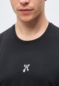 Κοντινό πλάνο στο κάτω μέρος του προσώπου και τον λαιμό ενός άνδρα που φοράει μαύρο μπλουζάκι με στρογγυλή λαιμόκοψη και μικρό λευκό αφηρημένο λογότυπο στο κέντρο του στήθους.