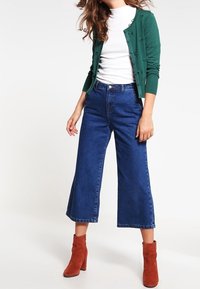 Cardigan verde lavorato a maglia con bottoni decorativi, t-shirt fitted bianca, jeans a vita alta e gamba larga blu, e stivaletti rossi alla caviglia.