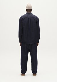 Donkerblauwe denim shirt en broek set met een relaxte pasvorm. Heeft lange mouwen en een detail op de rug. Draagt een beige gebreide muts.