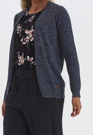 Femme portant un cardigan en maille bleu foncé sur une blouse noire à motifs floraux et un pantalon noir taille haute, devant un fond clair uni.