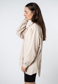 Licht beige langarmshirt van textuurstof, met knoopsluiting aan de voorkant en een oversized pasvorm. Iets langere achterkant, casual stijl.