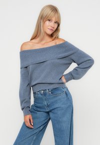 Εκτός ώμου πουλόβερ από μαλακό μπλε ύφασμα με ριγέ λαιμόκοψη, μακριά μανίκια και χαλαρή εφαρμογή, συνδυασμένο με τζιν παντελόνι ψηλής μέσης.