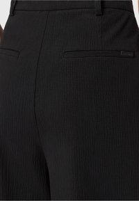 Close-up van zwarte gestreepte broek met riemlussen en twee achterzakken met paspel, waarvan er één een klein merkje heeft.