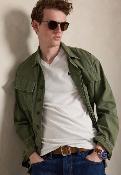 Mladý muž s okuliarmi na slnko, olivovo zelenou bundou cez bielu košeľu, modrými džínsami a hnedými koženými hodinkami, ruky v vreckách.