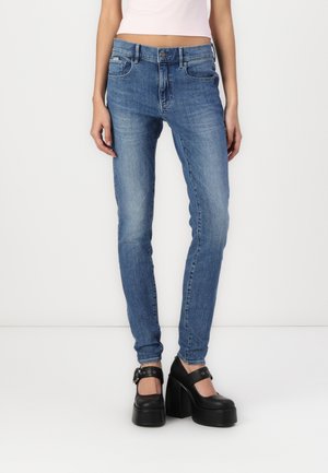 Femme portant un jean slim bleu délavé, un haut court rose clair et des chaussures Mary Jane à plateforme noires, debout sur un fond blanc.