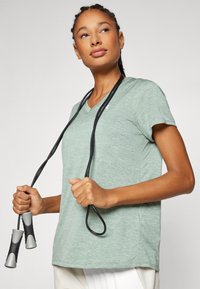 Camiseta deportiva de cuello en V verde claro, hecha de un tejido texturizado y transpirable. El modelo sostiene una cuerda de saltar con mangos negros y una cuerda enrollada.