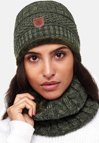 Cuffia lavorata a maglia verde oliva con trama a coste e fodera in pelliccia sintetica, con una toppa in pelle. Accompagnata da un passamontagna coordinato a coste.