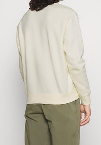 Světlé béžové mikina s kulatým výstřihem, žebrovanými manžetami a lemem a měkkou texturou. Nosí se s olivově zelenými kalhotami, pohled zobrazuje zadní design.