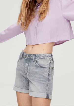 Jonge vrouw draag een lichtpaarse cropped blouse met knopen en opgerolde lichtgrijze denim shorts met een subtiele luipaardprint in de tailleband.