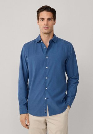 Chemise à manches longues, à boutons, en tissu bleu profond avec une finition texturée, présentant un col, un pli avant et un détail de marque sur le côté.
